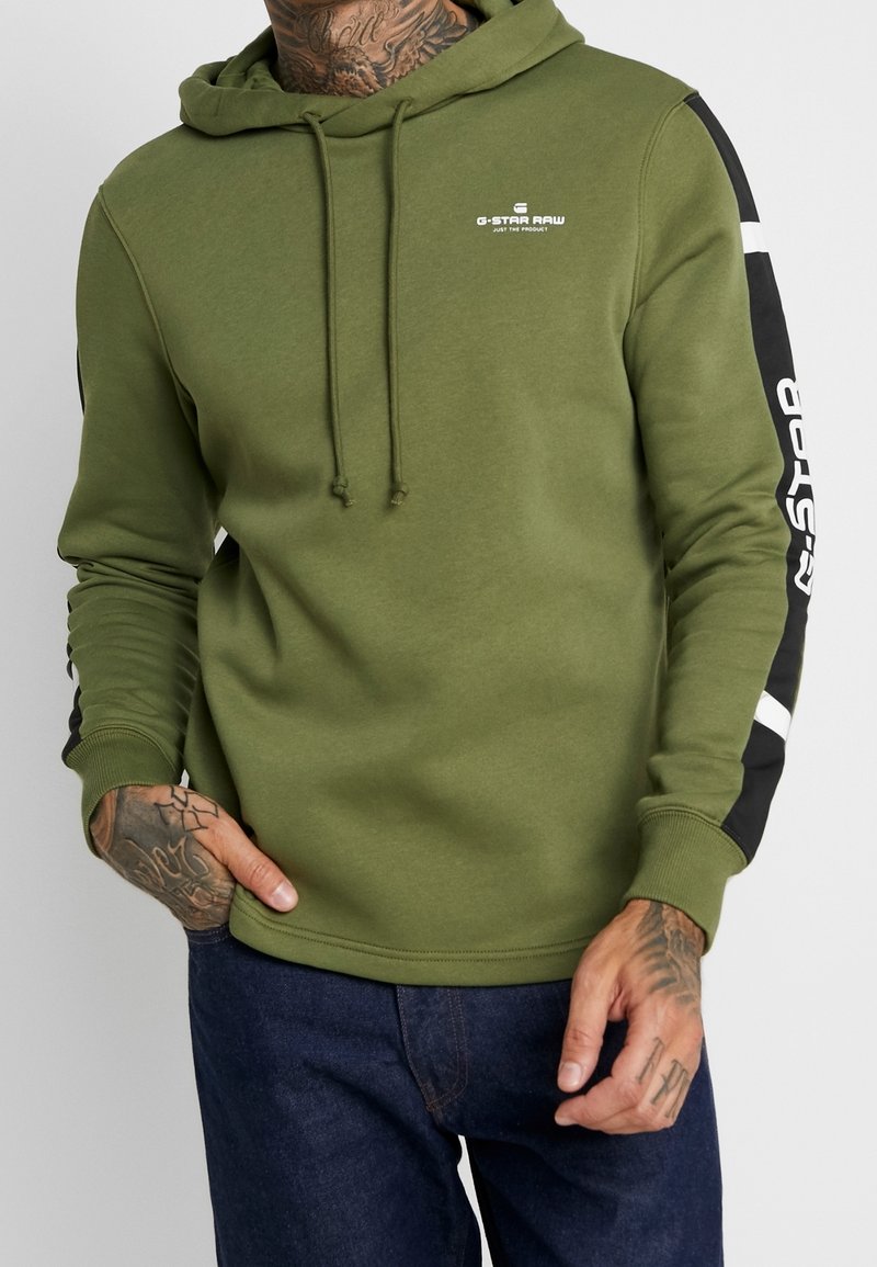 Olivgrön hoodie med en framficka i kängurustil, dragsko i huvan och svarta accenter på ärmarna med vit text. Texturerad tyg.