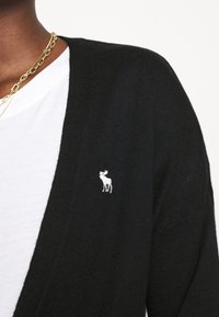 Persona vestida con un cárdigan negro con un pequeño logo de alce blanco sobre una camisa blanca, accessorios con dos cadenas de oro.
