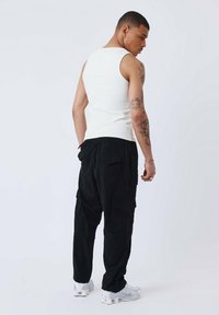 Camisetilla sin mangas blanca, pantalones cargo negros con bolsillos laterales y zapatillas blancas. Modelo visto desde atrás con tatuajes visibles en el brazo.