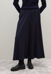 Navy wijde pijpen broek met een elastische tailleband, gemaakt van een gladde stof, gecombineerd met zwarte instappers en donkere sokken.