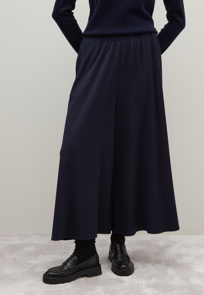 Navy wijde pijpen broek met een elastische tailleband, gemaakt van een gladde stof, gecombineerd met zwarte instappers en donkere sokken.