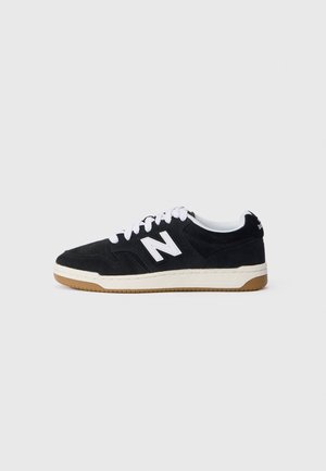 Czarny niski sneaker z białymi sznurowadłami i logo, beżowa podeszwa środkowa oraz gumowa podeszwa zewnętrzna na jednolitym jasnoszarym tle.