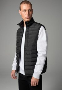 Chaleco negro acolchado con cuello alto, cremallera frontal y bolsillos laterales, superpuesto a una sudadera de color gris claro, contra un fondo neutro.