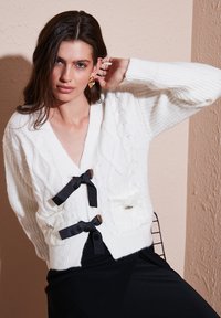Valge kudumitega cardigan mustade paeltega, V-kaelus, kaks esist pockets ja tekstuurne muster. Kombineeritud musta seelikuga.