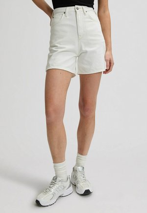 Persona che indossa pantaloncini di jeans bianchi a vita alta, calzini bianchi, scarpe sportive bianche e una maglietta nera, in piedi contro uno sfondo chiaro e uniforme.