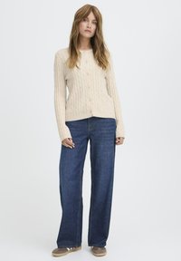 Beige kabelstrik cardigan med knapper foran, parret med løse mørk denim jeans. Model står med hænderne ned langs siden og neutrale sko.