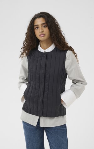 Mladá žena s dlouhými kudrnatými vlasy, oblečená v černé prošívané vestě přes světle šedé tričko a modré džíny, ruce v kapsách vesty.