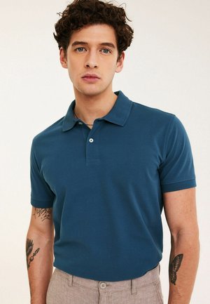Junger Mann mit lockigem Haar trägt ein blaues Poloshirt und beige Hosen, zeigt Tätowierungen eines Vogels und eines Schmetterlings auf seinen Unterarmen.