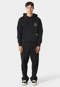 Czarna bluza z kapturem z kieszenią z przodu, z ściągaczem w kapturze i złotym nadrukiem. Połączona z luźnymi czarnymi spodniami i czarnymi trampkami z białymi sznurowadłami.