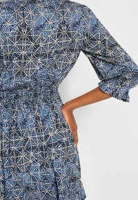 Abito a fantasia blu e bianco con vita aderente, caratterizzato da un design geometrico, maniche a campana e un tessuto strutturato.