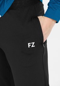 Person iført sorte bukser med FZ-logo, hånd placeret i den lynlåse side lomme, og en blå langærmet skjorte synlig ved håndleddet.