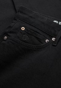 Tejido de mezclilla negra con un bolsillo frontal con un logo y remaches metálicos, textura suave y costuras detalladas.