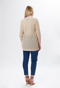 Cárdigan largo de punto beige con mangas de tres cuartos, que presenta un corte holgado y una textura ligeramente transparente, combinado con jeans azules.