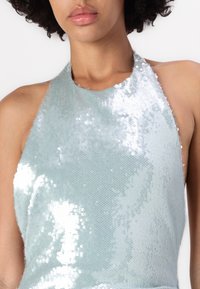 Ανοιχτό μπλε τοπ με λαιμόκοψη στο ύφος halter, φτιαγμένο από υφή καλυμμένη με γυαλιστερά, τετράγωνα παγιέτες, δημιουργώντας ένα κλιμακωτό εφέ από ματ σε αντανακλαστικό.