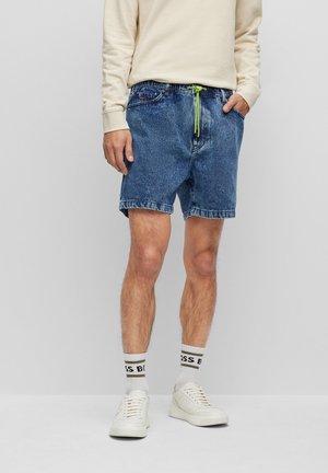 Uomo che indossa pantaloncini di jeans blu con coulisse verde, maglia a maniche lunghe color crema, calzini bianchi con scritte nere e scarpe da ginnastica bianche, in piedi davanti a uno sfondo grigio.
