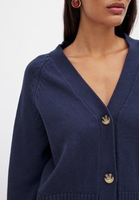 Granatowy kardigan z dekoltem w serek, o teksturowanym splocie i dwoma dużymi, okrągłymi guzikami. Posiada delikatne zdobienia na ramionach oraz ściągacz na dole.