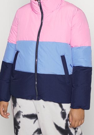 Kind draagt een roze, lichtblauwe en donkerblauwe pufferjas met gekleurde vlakken, een ritssluiting aan de voorkant en witte broek met zwarte tie-dye vlekken.