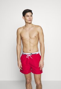 Rote Badeshorts aus schnelltrocknendem Material, mit einem weißen elastischen Bund mit schwarzem Branding und einem Kordelzug.