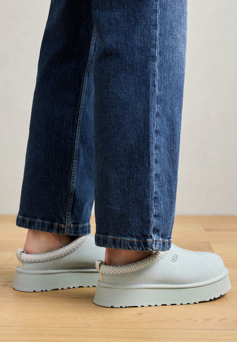 Hellgraue Slipper mit einer glatten Oberseite, texturiertem Kragen und dicker Sohle, kombiniert mit einer dunklen Jeans in einer holzverkleideten Innenumgebung.