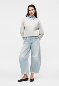 Vrouw met lang donker haar draagt een gelaagde grijze trui over een blauwe denim blouse, brede lichtgewassen cropped jeans met rafelige zoom en zwarte platte schoenen.