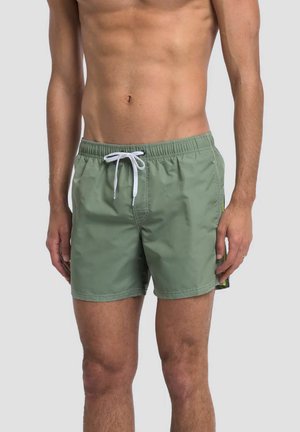 Uomo che indossa pantaloncini da bagno verde oliva con coulisse bianca, in piedi contro uno sfondo grigio chiaro uniforme.