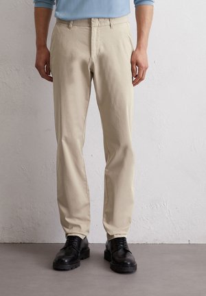 Beige Baumwollhosen mit geradem Schnitt, ausgestattet mit einem Reißverschluss und Taschen, kombiniert mit schwarzen Schnürschuhen auf einer grauen Oberfläche.