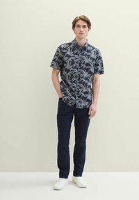 Kurzärmliges, navyblaues Hemd mit weißem Palmblattmuster, kombiniert mit dunkelblauen Hosen und weißen Sneakern, vor einem hellen Hintergrund stehend.