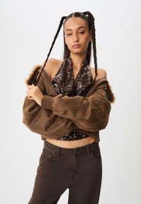 Hnedá skrátená bomber bunda s kožušinovým lemovaním cez trblietavé tričko, spárovaná s tmavozelenými priliehavými džínsami. Vlasy upravené do dvoch vrkôčikov.