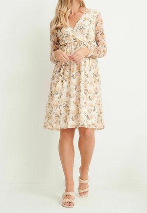Femme portant une robe fleurie à manches longues et longueur au genou, aux tons beiges et marron, assortie à des talons ouverts beige, debout devant un fond blanc.