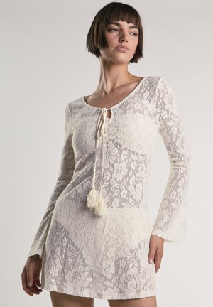 Donna che indossa un miniabito in pizzo crema a maniche lunghe con motivi floreali e lacci con nappine sullo scollo, posando davanti a uno sfondo neutro.