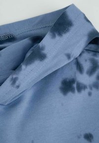 Niebieska tkanina o gładkiej fakturze z wzorem tie-dye w ciemniejszych odcieniach. Dekolt jest prążkowany, a szwy są widoczne wzdłuż krawędzi.