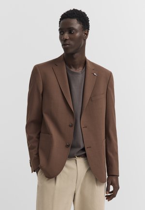 Ein Mann trägt einen braunen Blazer über einem dunkelgrauen T-Shirt und beigen Hosen, steht mit teilweise in den Taschen steckenden Händen und blickt zur Seite.
