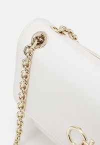 Bílá kožená kabelka s hladkou texturou, zlatým řetízkovým páskem a kruhovým kováním s vyraženou značkou.