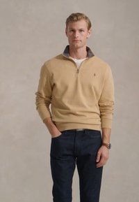 Šviesiai ruda zip-up megztinis su pilku apykakle, lengvas audinys, raukšlėti rankogaliai, derinamas su tamsiai mėlynomis džinsais. Nėra matomų išskirtinių raštų ar logotipų.