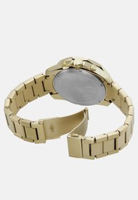 Kullatud toonis kell metallist rihmaga, millel on kindlad lingid, ümmargune korpus ja nupud külgedel. Tagakülg on sile ja roostevabast terasest.