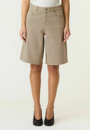 Kvinde iført beige knælange denimshorts kombineret med sorte højhælede sko, står mod en ensfarvet lys baggrund.