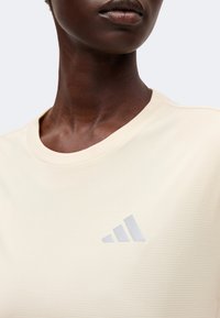 Gros plan d’une personne portant un t-shirt Adidas de couleur crème avec un petit logo Adidas argenté sur la poitrine.