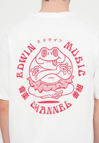 T-shirt branca de algodão com um gráfico vermelho de um sapo vestindo um tutuzinho com bolinhas, rodeado por texto circular que diz "EDWIN MUSIC CHANNEL."