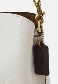 Bílá kožená taška s texturovaným povrchem, zlatými doplňky a hnědou koženou štítkou připevněnou řetízkem. Interiér je v kontrastní teplé hnědé barvě.