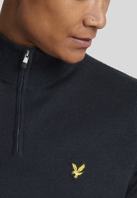 Navy knit zip-up sweater with a high collar, featuring a gold eagle logo on the chest and a smooth texture. Silver zipper detail.