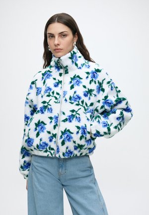 Kvinde iført en hvid fleecejakke med blåt blomsterprint og grønne blade, kombineret med lyseblå jeans, stående med den ene hånd i lommen.