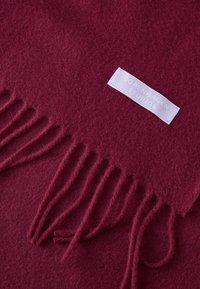 Bufanda de lana burdeos con bordes con flecos y una etiqueta blanca con el texto "Copenhagen Studios". Textura suave y acabado de tela uniforme.