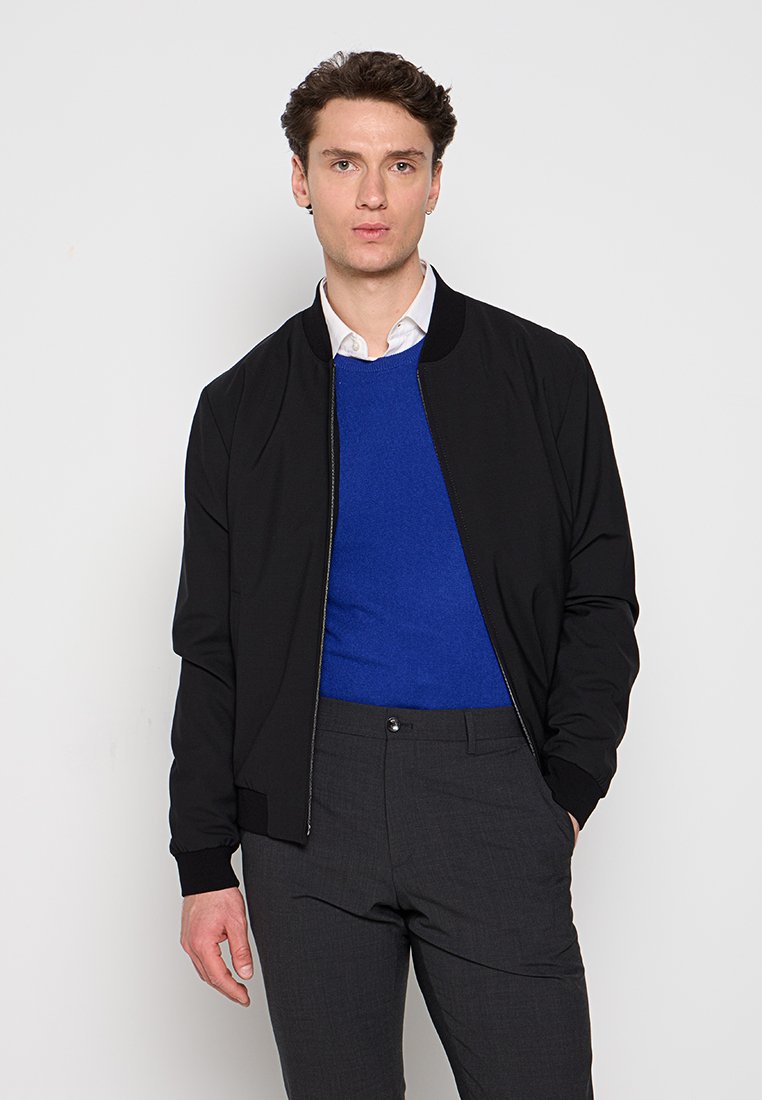 Schwarze Bomberjacke mit Reißverschluss, gerippten Bündchen, getragen über einem blauen Pullover und grauen Hosen. Glattes Material und minimalistisches Design.