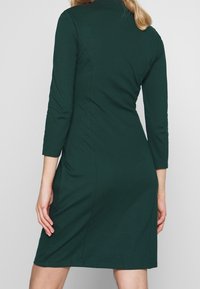 Femme portant une robe ajustée vert foncé longueur genou avec des manches trois-quarts, vue de dos sur un fond uni.