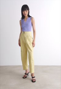 Top sans manches lilas avec un col en V ; pantalon jaune taille haute avec des plis et des poches ; sandales à talons noires. Fond uni.
