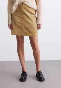 Jupe en velours côtelé kaki avec une coupe droite, poche avant et fermeture à boutons, associée à des chaussures noires à enfiler. Pull texturé de couleur neutre.