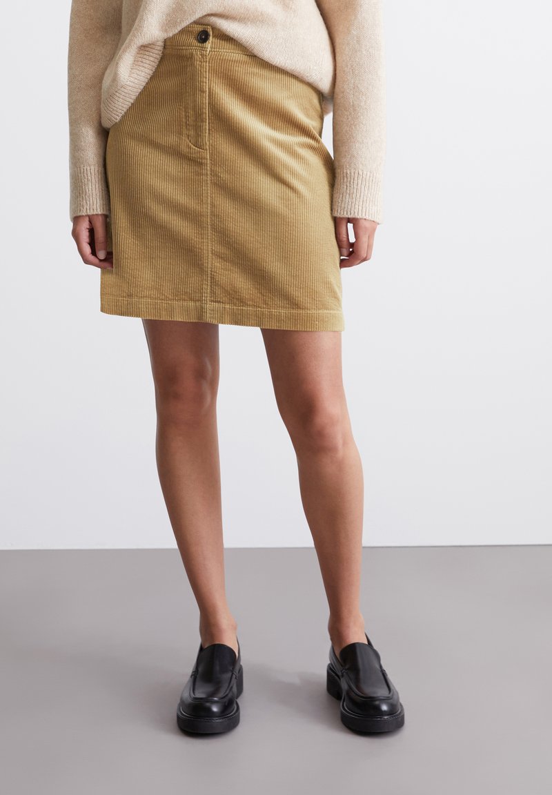 Jupe en velours côtelé kaki avec une coupe droite, poche avant et fermeture à boutons, associée à des chaussures noires à enfiler. Pull texturé de couleur neutre.