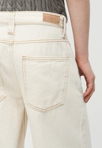 Άτομο που φοράει κρεμ τζιν από ντένιμ και γκρι πλεκτό τοπ, δείχνοντας την πίσω τσέπη και την δερμάτινη ετικέτα της μάρκας στη μέση.