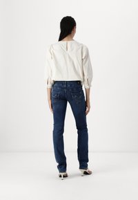 Blusa blanca de manga larga con mangas abullonadas y escote en la espalda, combinada con jeans de mezclilla azul oscuro que tienen bolsillos traseros y detalles de costura.