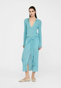 Šviesiai mėlyna suknelė su briaunotu audiniu, gilias V formos iškirpte, ilgomis rankovėmis ir juosmens raišteliu, su šonine skeltuke ir iki kelių siekiančia apačia. Dėvima su šviesiais batais.